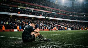 A cinematic, high-contrast editorial shot of a dejected professional footballer sitting alone on a r